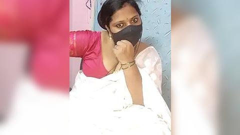 Media: Video of a South Asian woman in a pink blouse, black face mask, and white sari, lying on a bed with a blue wall behind her, blurred background.