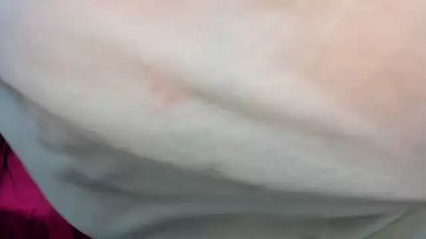 Media: Video of a close-up of a person's upper back, with a noticeable bruise on the left side. The background is blurred, featuring a pinkish fabric. The skin tone appears pale, and the lighting is soft.