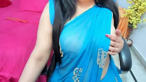 Media: Video of a woman in a bright blue sari, revealing her left breast through the sheer fabric. She has long black hair and is seated on a red bed.