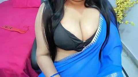 Media: Video of a South Asian woman with long black hair, wearing a black bra and a blue saree, seated on a pink bed, with yellow flowers in the background.