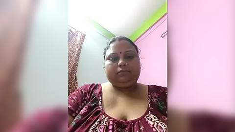 Media: A video of a plus-size, dark-skinned woman with a round face, wearing a floral maroon top, standing in a pink-painted room with green ceiling.