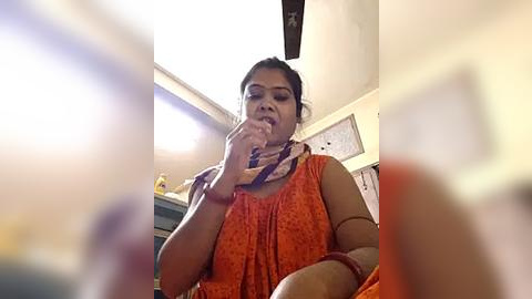 Media: Video of a woman in an orange sari, talking on a phone, in a dimly lit room with a ceiling fan, cluttered background.