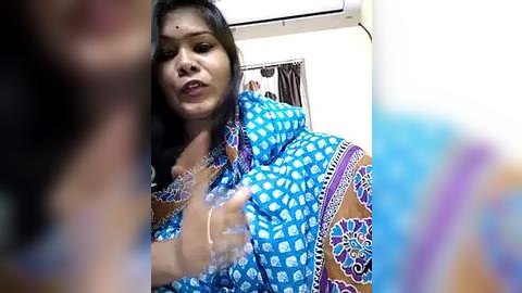 Media: Video of a South Asian woman with long black hair, wearing a blue sari with intricate white floral patterns, gesturing with her right hand. Background features a white wall and a ceiling-mounted air conditioner.