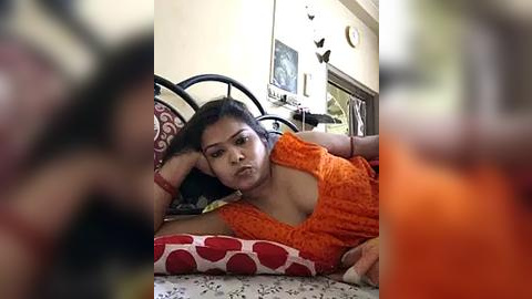 Media: A video shows an Indian woman with medium skin tone, wearing an orange patterned top, lying on a bed with red polka-dot pillows. The background includes a clock, a mirror, and a cluttered room.