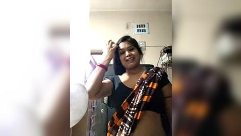 Media: A video of a smiling South Asian woman with dark hair, wearing a black top and a colorful sari, adjusting her hair in a domestic room with a white fan and cluttered background.