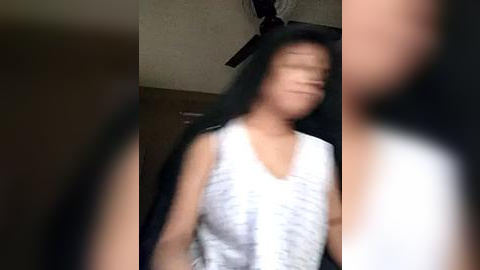 Media: A blurry video of a woman with long black hair, wearing a sleeveless white top, standing in a dimly lit room with a ceiling fan. The background includes dark, possibly wooden, furniture.