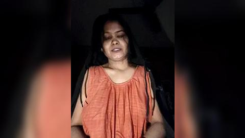 Media: Video of a middle-aged South Asian woman with medium skin tone and long black hair, wearing an orange sleeveless blouse, seated in a dimly lit room with shadows.