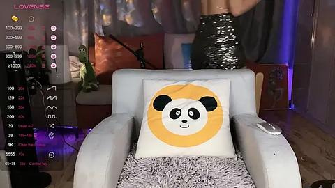 Media: Video of a cozy living room with a plush grey armchair featuring a decorative pillow with a panda face. The background includes a plush orange cushion, a potted plant, and a person partially visible in a shiny, black sequined dress.