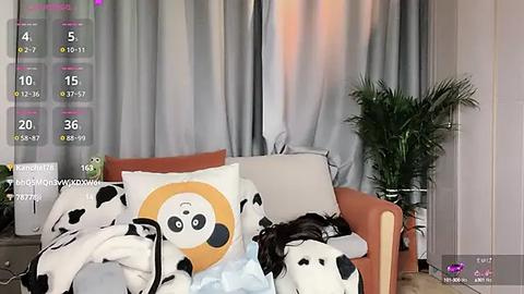 Media: Video of a woman with dark hair, wearing a panda-patterned blanket, lying on a beige couch with a potted plant and gray curtains in the background.