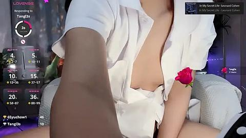 Media: Video of a partially dressed woman with fair skin, wearing a white button-up shirt, revealing cleavage, sitting on a couch with a red rose in her hair.