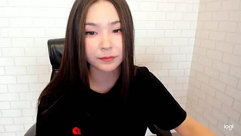 Media: Video of an East Asian woman with long black hair, fair skin, and a slight smile, wearing a black T-shirt, seated indoors against a white brick wall.