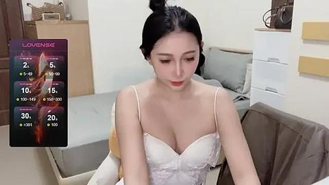 Media: Video of an East Asian woman with fair skin and black hair tied up, wearing a white lace bra, sitting in a modern bedroom with beige furniture.