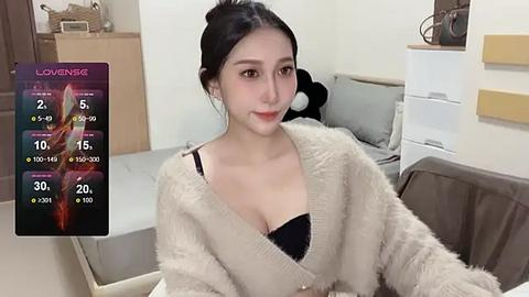 Media: Video of an Asian woman with fair skin, black hair in a bun, wearing a beige fuzzy cardigan over a black bra. Background shows a neatly made bed, white wall, and wooden furniture.