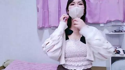 Media: Video of an Asian woman with a pale complexion, long black hair, wearing a white face mask, white jacket, polka-dot blouse, and adjusting her mask in a pink-draped room.