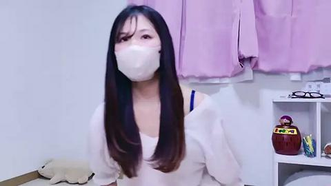 Media: Video of an East Asian woman with long black hair, wearing a white mask and shirt, sitting in a room with pastel pink curtains, a stuffed toy, and a red box.