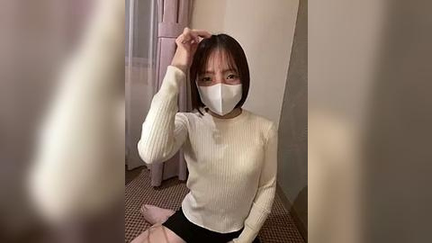 Media: Video of an East Asian woman with straight black hair and bangs, wearing a white ribbed sweater, black skirt, and face mask, sitting on a beige carpet, touching her hair.