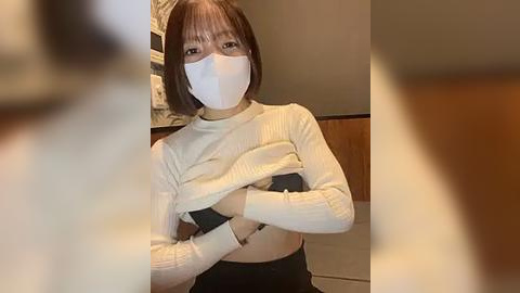 Media: Video of an Asian woman with a short bob haircut, wearing a white face mask, white sweater, and black pants, in an indoor setting with blurred background.