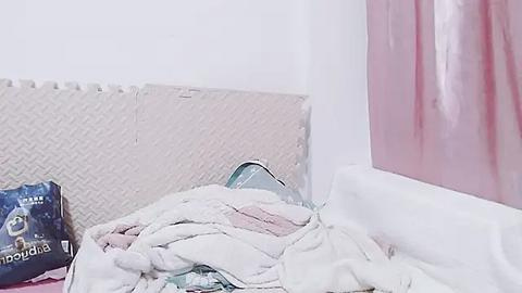 Media: Video of a cluttered room with a beige rug, a partially visible pink curtain, and a disorganized pile of blankets and pillows on the floor.