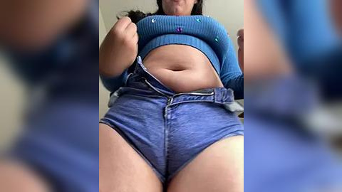 Media: Video of a woman lifting her blue denim shorts, revealing a midriff and blue crop top, taken from a low angle.