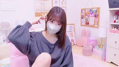 Media: Video of an Asian woman in a gray sweatshirt, wearing a face mask, sitting on a pink chair in a pastel-colored, bunny-themed bedroom with framed artwork, soft lighting, and plush toys.