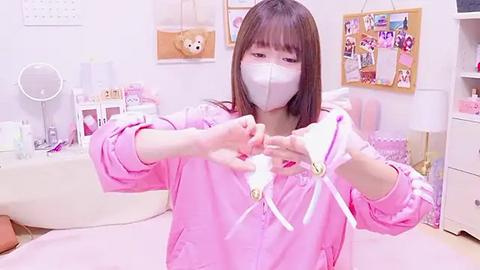 Media: Video of an East Asian woman in a pink surgical mask, wearing a pink robe, preparing white gloves with yellow tips in a pastel-toned, decorated room.