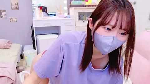 Media: Video of an Asian woman with straight, shoulder-length brown hair and pale skin, wearing a light blue surgical mask, light blue scrubs, and a white cap, in a cluttered, brightly lit medical office with pastel-colored walls and shelves.