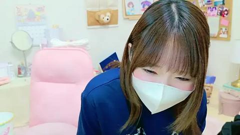 Media: Video of an Asian woman with long brown hair, wearing a blue medical gown, and a white mask, sitting in a pink chair in a brightly lit, child-friendly clinic.