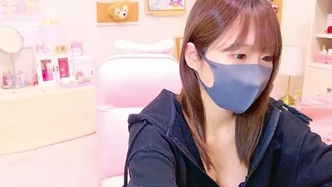 Media: Video of a young Asian woman with straight, shoulder-length brown hair, wearing a blue face mask and black hoodie, seated in a brightly lit, pastel-themed salon with pink walls and various beauty products on a counter.