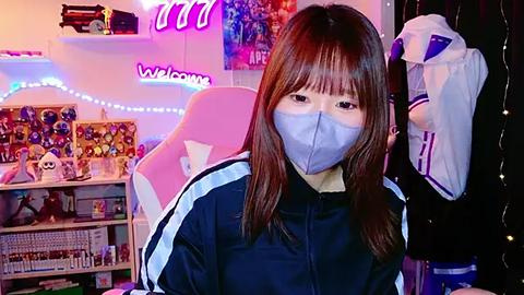 Media: Video of an East Asian woman with long brown hair, wearing a blue face mask and black tracksuit, sitting in a pink gaming chair in a colorful, cluttered bedroom with gaming posters and plushies.