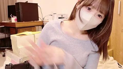 Media: A video of a young Asian woman with long brown hair, wearing a light grey sweater, white mask, and gloves, sitting in a cluttered office with a printer, chair, and papers.