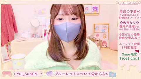 Media: A video of a young woman with straight brown hair and light skin, wearing a light green sweater and blue face mask, seated in a cozy room with light pink walls and framed pictures.