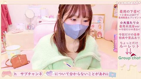 Media: A soft-focus video of an Asian woman with light skin and long brown hair, wearing a blue mask and green sweater, in a pastel-colored room with Japanese text and a pink cat figurine.