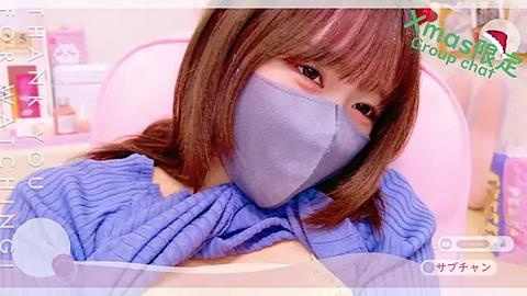 Media: A close-up video of a young woman with straight brown hair, wearing a blue face mask and blue knit top, reclining on a pink chair in a softly lit room.