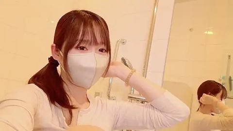 Media: Video of an East Asian woman with straight brown hair, wearing a white mask and white top, standing in a modern bathroom with white tiles and a showerhead.