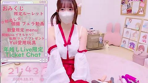 Media: A video of a young Japanese woman in a white kimono with red accents, wearing a face mask, sitting in a pink room with anime character posters and a table.