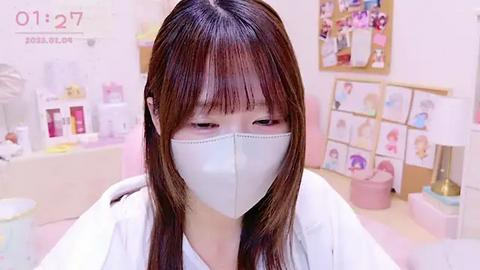 Media: Video of a young Asian woman with long, straight brown hair, wearing a white mask and lab coat, standing in a brightly lit, colorful art studio with pastel walls, art supplies, and a bulletin board.