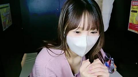 Media: A video shows an Asian woman with long brown hair and a white mask, sitting indoors, wearing a pink sweater, and holding her hands together.