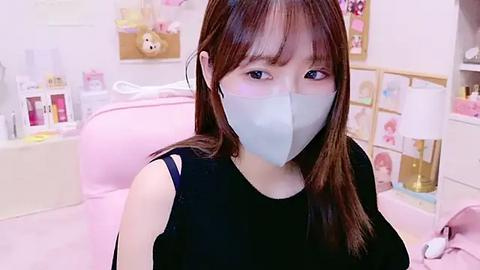 Media: Video of an East Asian woman with long brown hair and light skin, wearing a black sleeveless top, gray mask, and headphones, in a pink bedroom with white furniture and stuffed animals.