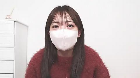 Media: A video of an East Asian woman with long black hair, wearing a white face mask and a red fuzzy sweater, standing in a plain white room with a white dresser in the background.