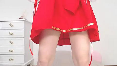 Media: Video of a woman in a red, garter-belted skirt, standing in a bedroom with a white dresser and a pink cushion in the background.