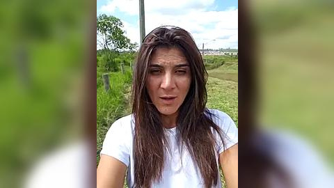 Media: Video of a young woman with long brown hair, wearing a white T-shirt, standing outdoors on a grassy field with a clear sky and distant trees.