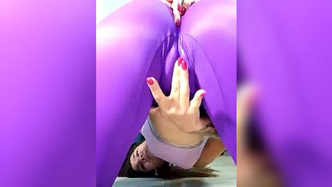 Media: Video of a woman with long brown hair and red nails, wearing purple pants, lying on a bed, reaching out to touch her exposed vagina.