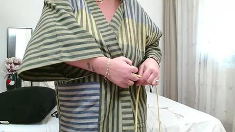 Media: A video of a person in a striped, green and blue robe, adjusting a yellow cord in a softly lit, tidy bedroom with white curtains and a bed.