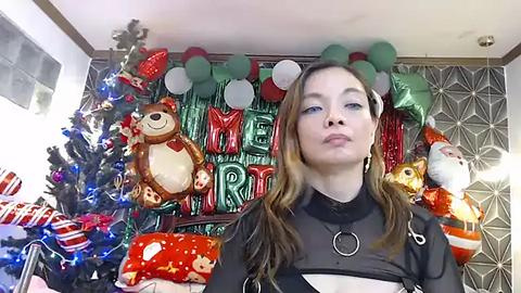 Media: Video of a young woman with long brown hair, wearing a black top, standing in front of a festive Christmas tree adorned with red and green decorations and a \"Merry Christmas\" banner.