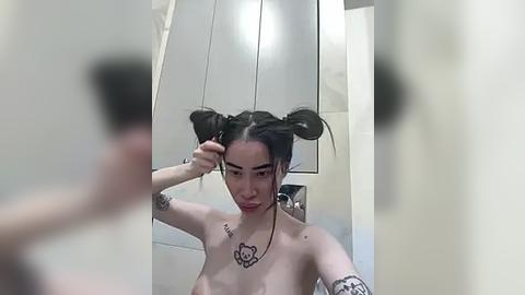 Media: Video of a young, fair-skinned woman with dark hair in two high pigtails, topless, with a rose tattoo on her chest, standing in a bathroom, adjusting her hair.