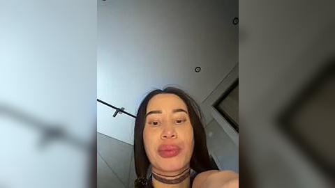 Media: Video of a young woman with long black hair and fair skin, wearing a black choker, puckering her lips, in a modern, minimalist room with a white ceiling and exposed light fixtures.