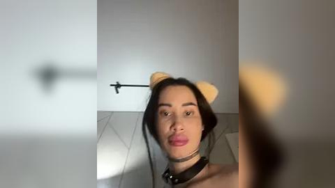 Media: Video of a young woman with fair skin, brown hair, wearing a black choker and cat ears, smiling in a modern, minimalist room with light-colored walls and tiled floor.