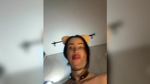 Media: Video of a woman with fair skin, dark hair, and red lipstick, wearing a black choker and a dog headband, in a modern bathroom with a white wall and chrome towel rack.