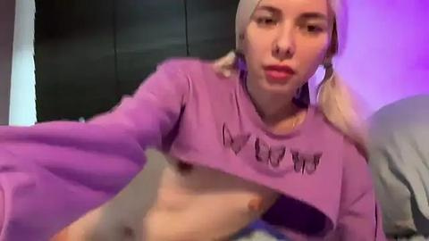 Media: Video of a young woman with pale skin, blonde pigtails, and a purple crop top exposing her small breasts. She's indoors, on a bed with grey sheets, in a dimly lit room with purple lighting.