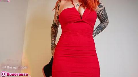 Media: Video of a fair-skinned woman with long red hair wearing a tight, strapless red dress that accentuates her ample cleavage. Her arms are adorned with detailed black tribal tattoos. She stands against a plain, light-colored background.